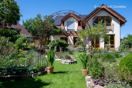 Blick über einen Garten mit Rosenbogen und blühenden Beeten auf ein hell verputztes Haus mit hellbraunen Fensterrahmen