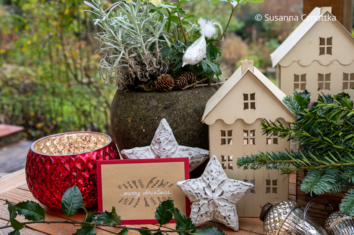 Weihnachtsdekoration auf dem Terrassentisch Weihnachtsdekoration auf dem Terrassentisch mit weißen Metallhäuschen, rotem Windlicht und Karte mit dem Schriftzug "Merry Christmas"