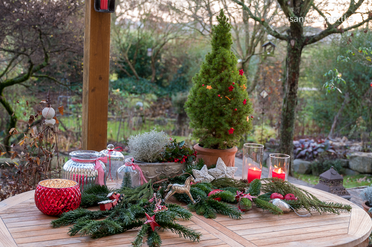 Ein Terrassentisch ist mit weihnachtlichem Grün, Deko in Weiß und Silber und roten Kerzen geschmückt