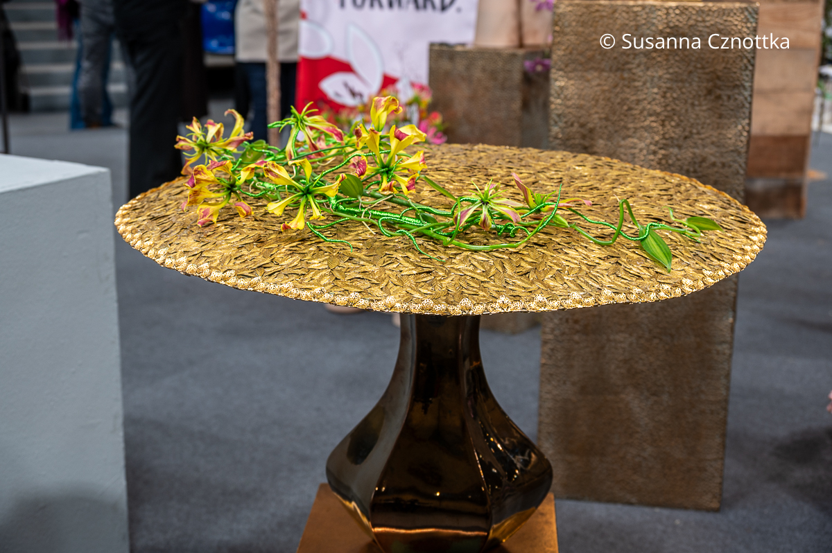 Floristik auf der IPM Essen: eine runde  Basis mit golden gefärbten Blüten von Plattährengras mit wenigen Stielen der Ruhmeskrone