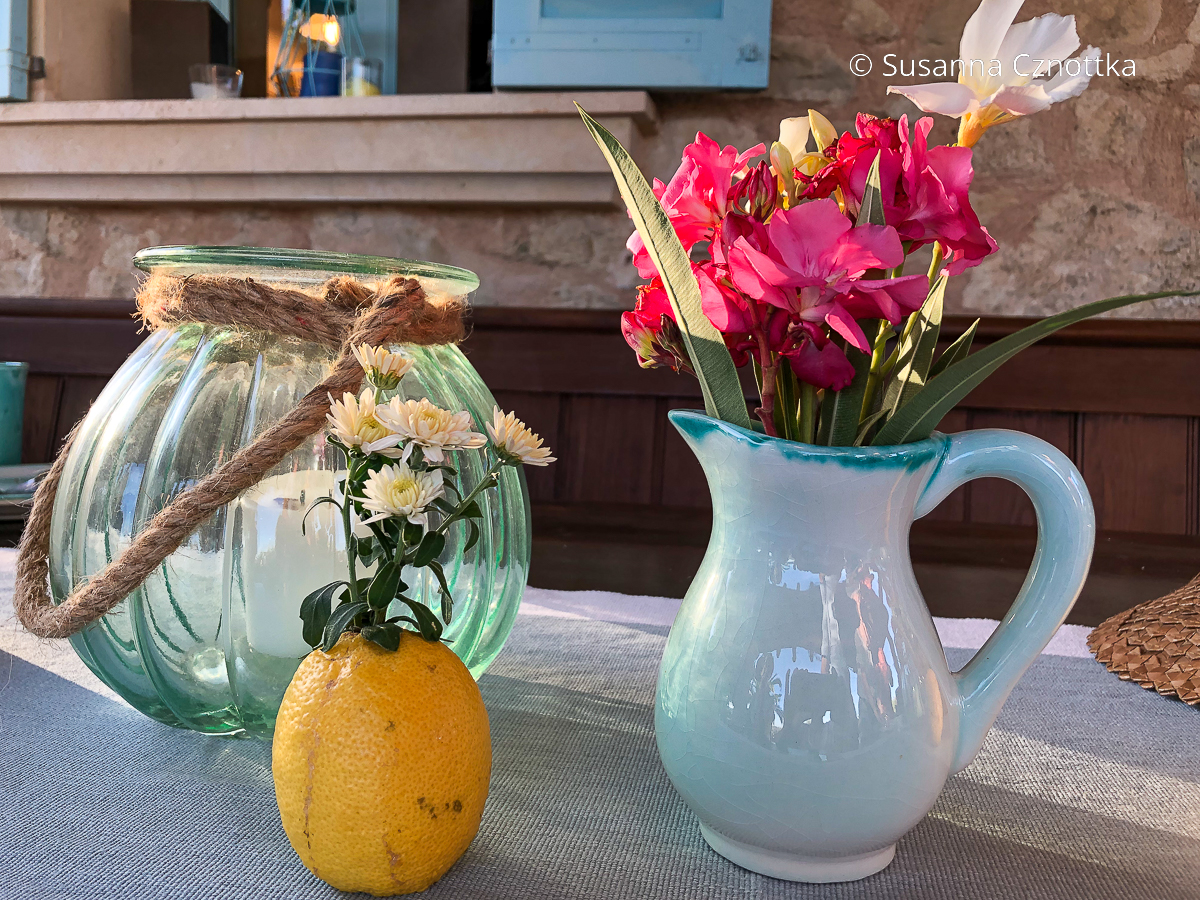Eine Vase mit Blumen, ein Windlicht und und eine Zitrone mit Blüten darin schmücken einen Tisch