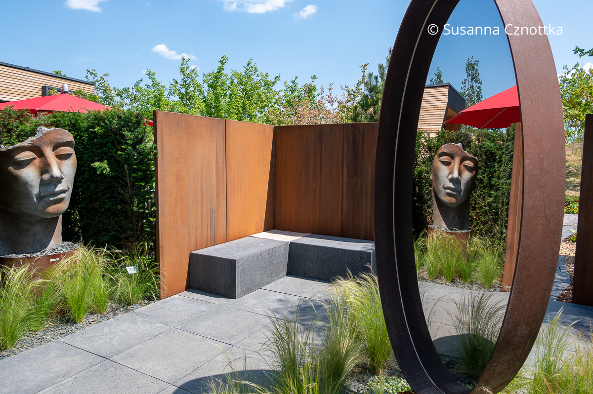 Die Skulptur eines Gesichts spiegelt sich in einem drehbaren Spiegel in einem Schaugarten auf der BUGA Mannheim 2023