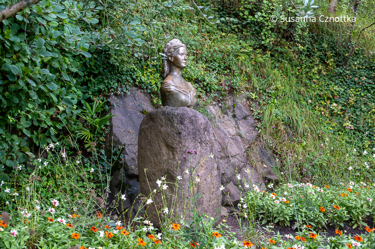 Eine Büste der Kaiserin Sissi auf einem großen Stein, davor weiße und orange Blumen