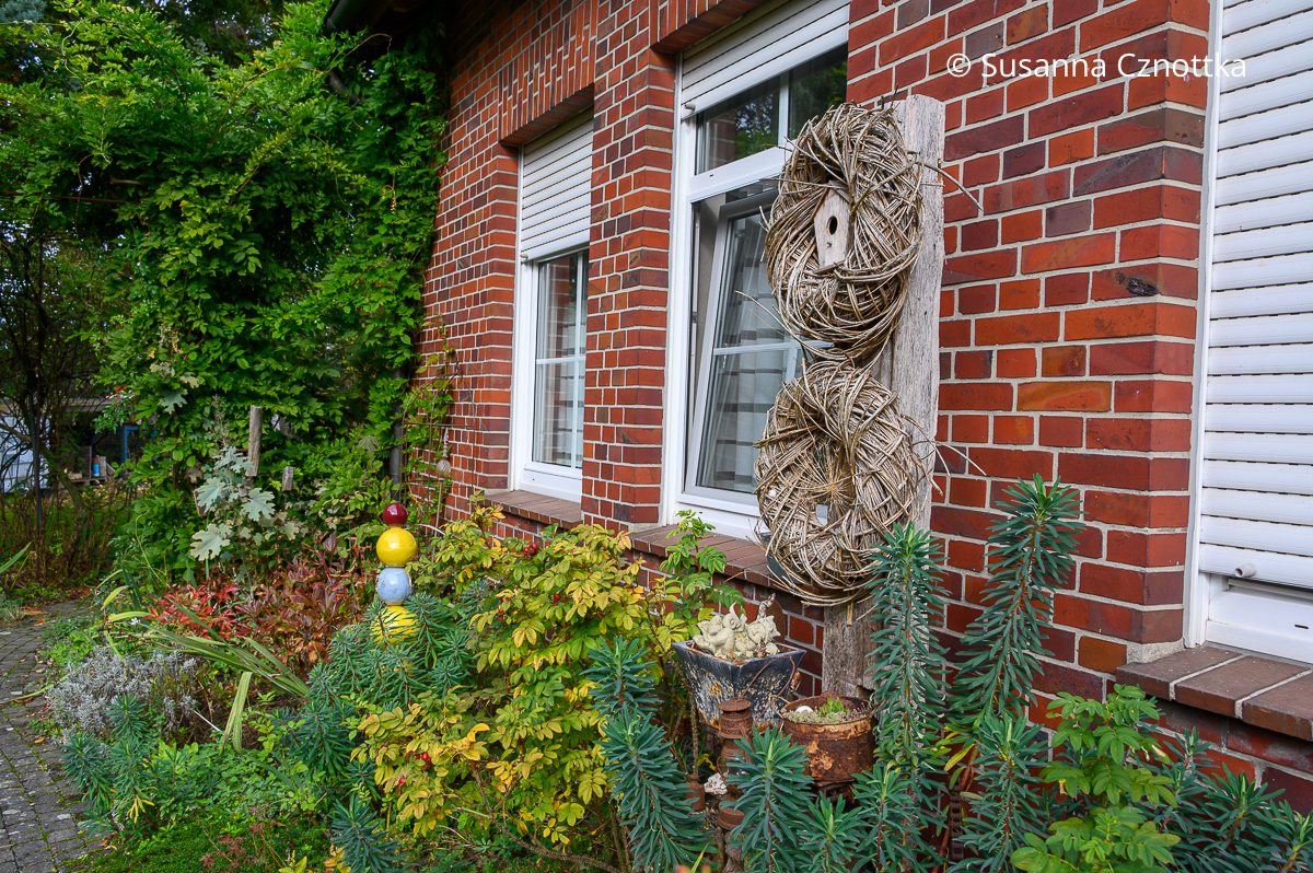 Verschiedene Laubfarben und zwei schlichte Kränze an einem aufrechten Balken an der Rückseite eines Hauses aus rotem Klinker