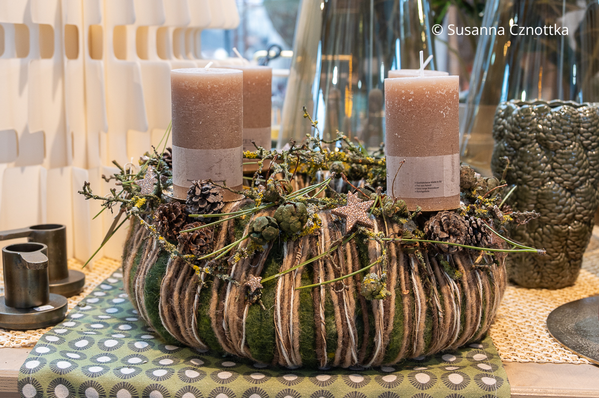 Adventskranz auf einer Filzunterlage mit vier dicken beigen Kerzen, Bändern in Naturfarben und kleinen Zapfen und Sternen