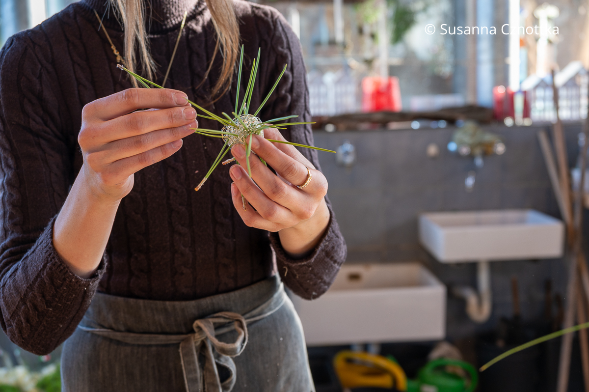 Eine junge Frau steckt Piniennadeln in eine Kugel aus dünnem silbernem Draht