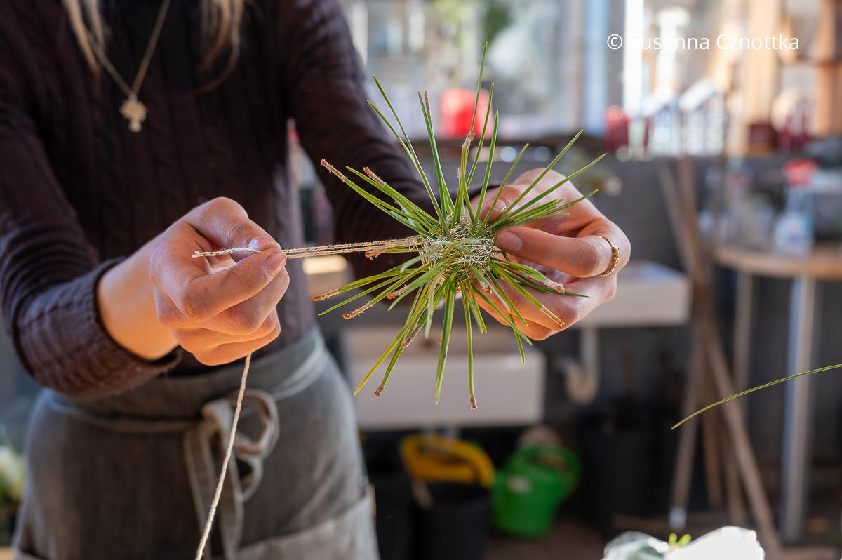 Eine junge Frau befestigt eine Schnur zum Aufhängen an einer Kugel aus silbernem Draht und Piniennadeln