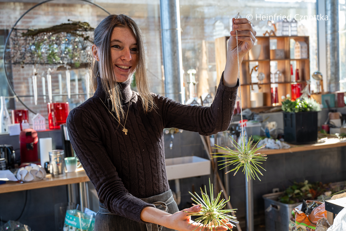 Eine junge Frau zeigt zwei fertige Sterne aus silbernem Draht und Piniennadeln