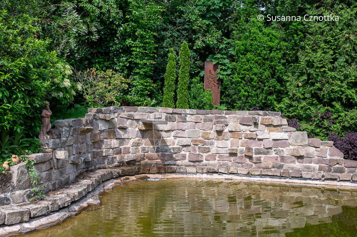 Blick über einen Schwimmteich mit Natursteinmauer und Wasserspiel