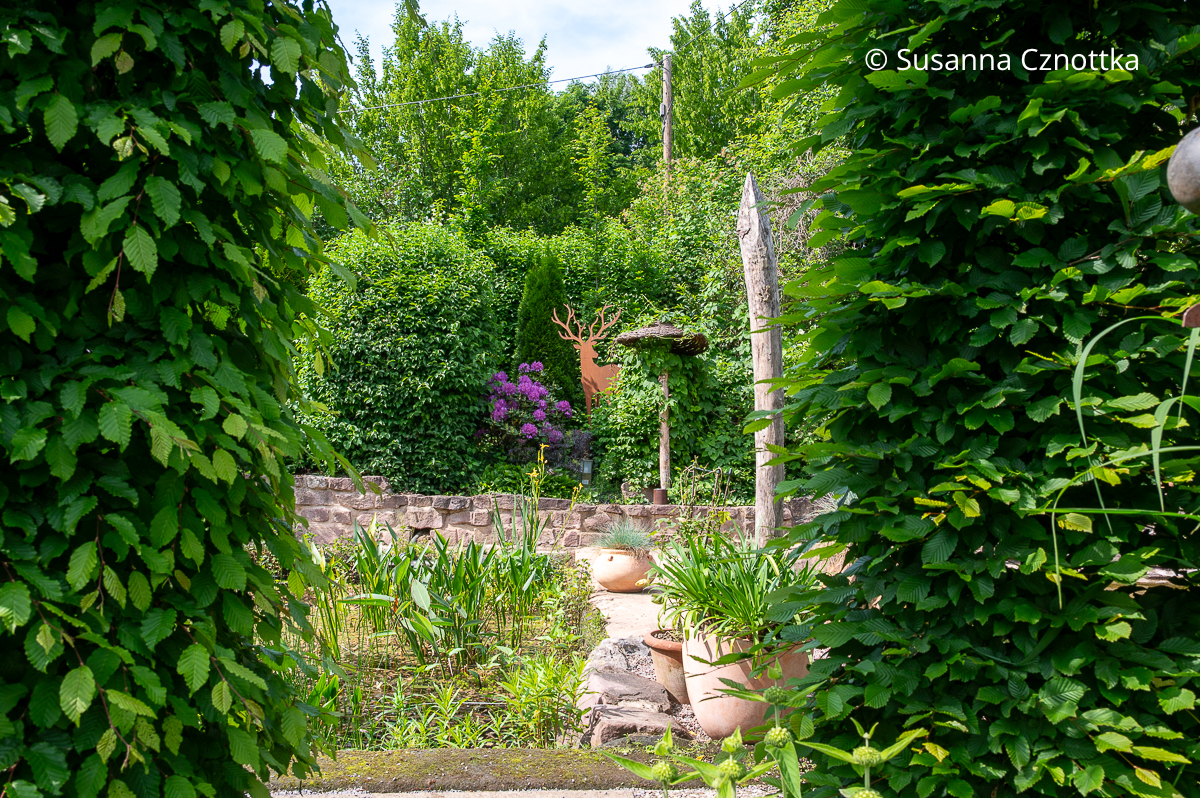 Blickachse: blühender Rhododendron und Gartendeko zwischen zwei Säulen aus Hainbuche