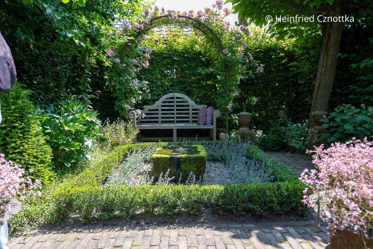 Ein kleiner formaler Garten mit einem Quellstein, rechteckigem mit Eibe eingefassten Beet und einer Lutyens-Bank unter einem Rosenbogen Ein kleiner formaler Garten mit einem Quellstein, rechteckigem mit Eibe eingefassten Beet und einer Lutyens-Bank unter einem Rosenbogen mit einer rosa Kletterrose